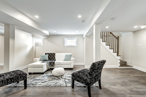 A beautifully remodeled basement. The decor is mostly white with wood floors. Chairs, a sofa, and modern carpet also adorn the space.